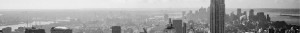 Panoramic black-and-white view of a city skyline with numerous buildings of varying heights. The tallest skyscraper on the right, possibly housing government work, dominates the scene. The background shows a hazy horizon under a partly cloudy sky, with bridges and rivers visible.