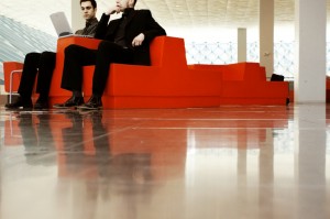 Two men wearing suits sit on modern, red, geometric chairs with a laptop in an open, brightly lit space that features reflective flooring and a large window with a blue grid pattern in the background. The setting appears to be a contemporary workspace or lobby, perfect for managing people effectively.