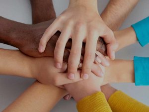 A diverse group of hands stacked together in a united gesture. The hands belong to people of different races and genders, symbolizing teamwork, cooperation, and solidarity.