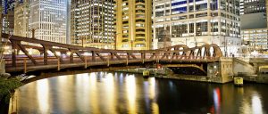 A night view of a city skyline with tall buildings illuminating the scene. In the foreground, a lit-up bridge spans a calm river, reflecting the lights from the structures. The architecture combines modern glass facades and classic designs.