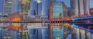 City skyline at dusk reflected in the water. Buildings of varying heights and styles, including glass skyscrapers and brick structures, are illuminated with golden and blue lights. A bridge with blue lights spans the right side, creating a vivid, mirrored effect.