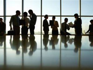 Silhouetted group of people standing and conversing in front of large, bright windows. They appear to be in an office setting, with some holding laptops and others engaged in discussion. The reflective surface of a table is visible in the foreground.