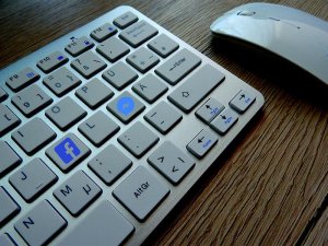 Close-up of a keyboard and mouse on a wooden surface. The keyboard has standard QWERTY keys along with a Facebook key replacing the "Ctrl" key and a Messenger key replacing the "Windows" key. The mouse is sleek and white.