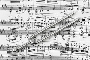 A tuning fork lies on top of a sheet of music. The sheet music is densely covered with various musical notes and symbols. The tuning fork, a metallic instrument with two prongs, is centrally placed on the sheet, underscoring its essential role in facilitating accurate pitches for public agencies and musicians alike.