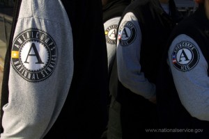 Close-up of three people standing side by side, each wearing a jacket with the AmeriCorps logo on the sleeve. The prominent "A" and "AmeriCorps National Service" are clearly visible. The background is blurred, while a website address can be seen at the bottom right.