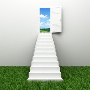 A white staircase rises from a grassy field, leading to an open door in a wall. The door reveals a bright blue sky with fluffy white clouds and a scenic landscape in the background. The image evokes a sense of transcendence and opportunity, much like embarking on the journey to your dream job.