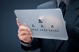 A person in a suit holding a transparent tablet displaying a hierarchical organizational chart. The chart shows a top leader connected to sub-leaders and further subordinate members, illustrating a structured chain of command, common among IT professionals.