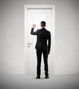 A person in a black suit is shown from behind, knocking on a white door. The individual, likely heading to an interview, has short brown hair and stands in a minimalist, gray-toned room with no visible decorations.