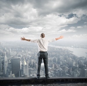 A leader in business attire stands on the edge of a rooftop with his arms outstretched, overlooking a sprawling cityscape under a cloudy sky.