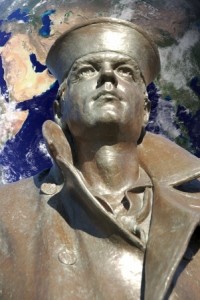 A bronze statue of a sailor, donning a cap and a coat with a large collar, looking upward. In the background, an image of Earth with visible continents and oceans underscores a global military theme.