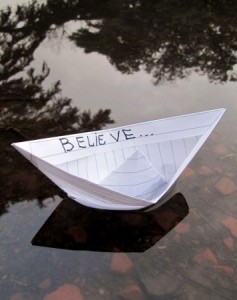 A small paper boat floats on water, reflecting an overcast sky and blurred trees. Crafted from lined notebook paper, it bears the word "BELIEVE" on one side—a simple reminder that even a public service career can start with a humble beginning.