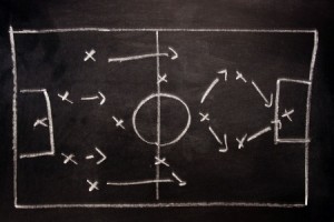 A chalkboard drawing of a soccer field with goalposts. The field is divided in half with a large circle in the center. Various "X" and arrow markings illustrate player positions and movement strategies on both sides, emphasizing performance goals for effective gameplay.