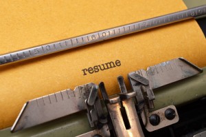 A close-up of a typewriter typing the word "resume writing" on a sheet of yellow paper. The metal type arms and the ruler bar are visible. The paper is slightly textured, and the typewriter is an older model with visible mechanical parts.