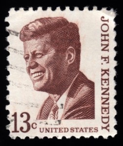 A vintage U.S. postage stamp featuring a detailed engraved portrait of John F. Kennedy in brown ink. “JOHN F. KENNEDY” is written vertically on the right side, and “13c UNITED STATES” is at the bottom, honoring his legacy of public service.