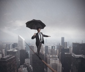A man in a business suit balances on a tightrope high above a city skyline on a rainy day, illustrating the precarious nature of government work. He holds an open black umbrella and extends his other arm for balance, with city buildings and a cloudy sky visible in the background.