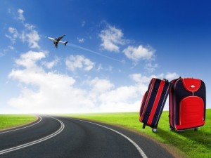 Two suitcases, one upright and one leaning against it, are placed on a green grassy area next to an asphalt road with a dashed white line. In the background, a plane is flying in a clear blue sky with several fluffy clouds, capturing the essence of travel adventures.