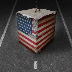 A large, weathered concrete block, partially covered with the American flag, sits on a dark asphalt road. The block appears to be tethered by an old, rusted metal hook protruding from its top. White dashed lines run parallel on either side of the block, evoking a sense of government authority.
