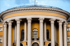 A neoclassical building with a yellow facade features six tall, fluted columns supporting a semi-circular portico. The columns have ornate capitals, and the structure includes multiple arched windows. A decorative railing tops the building against a clear blue sky, evoking an air of authority often associated with government managers.