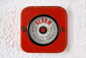 Close-up of a red emergency alarm button on a white wall. The alarm, clearly vital for an Emergency Manager, instructs users to break the glass to activate it, including the phrase "SMASH GLASS" in French, German, and Italian. The button has a weathered, slightly rusted appearance.