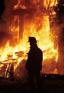 Silhouette of a firefighter wearing a helmet and standing resolutely in front of a massive blaze, with flames consuming a structure. The fire illuminates the night, highlighting the firefighter's stance.
