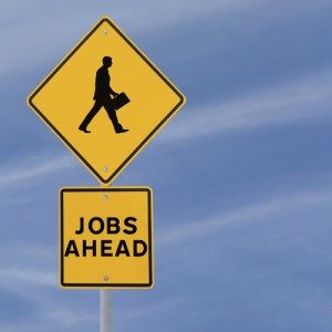 A yellow diamond-shaped road sign features the silhouette of a walking person carrying a briefcase. Below it, a rectangular yellow sign reads "JOBS AHEAD" in bold black letters. The word "Job" stands out prominently against the clear blue sky with faint wispy clouds.