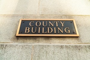 A close-up view of a sign mounted on a stone wall, displaying the words "COUNTY BUILDING" in uppercase letters. The sign is rectangular with a dark background and a gold-colored border and text, symbolizing the importance of local government.