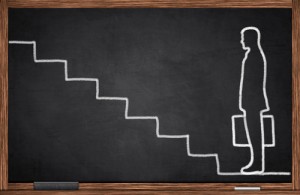 A chalk drawing on a blackboard depicts a person with a briefcase standing beside a series of ascending steps, symbolizing promotion. The wooden frame of the blackboard is visible around the edges, emphasizing the journey towards progress.