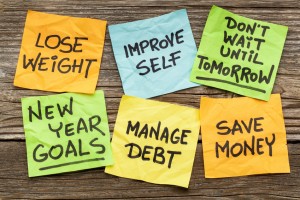 Six colorful sticky notes on a wooden surface display motivational messages for your resolutions: "LOSE WEIGHT," "IMPROVE SELF," "DON'T WAIT UNTIL TOMORROW," "NEW YEAR GOALS," "MANAGE DEBT," and "SAVE MONEY.