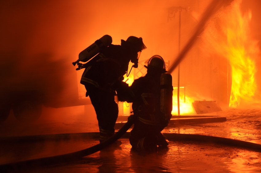 Two firefighters in protective gear and breathing apparatus work together to extinguish a blazing fire. The scene is engulfed in bright orange flames and thick smoke, with water hoses spraying in an intense firefighter effort.