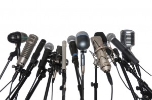 A collection of various types of microphones, including dynamic, condenser, ribbon, and handheld models, is arranged on adjustable microphone stands against a white background. The mics are positioned close together in a semi-circle formation, perfect for public speaking engagements.