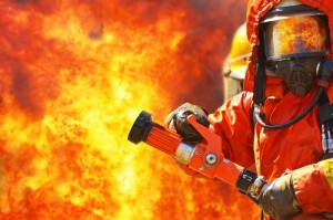 A firefighter in full protective gear, including a helmet and reflective visor, aims a fire hose against a backdrop of intense flames. Clad in a bright orange suit that emphasizes the heat and intensity of the blaze, this brave member of the fire service battles the inferno with unwavering resolve.