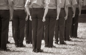 A sepia-toned image of job seekers standing in formation, seen from behind. They are wearing uniforms with hands clenched into fists hanging by their sides, and are standing on a grassy field.
