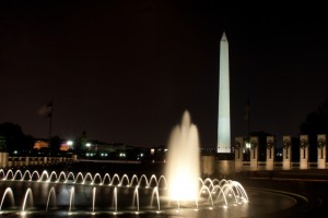 A nighttime view of the Washington Monument illuminated against the dark sky. In the foreground, a circular fountain with white streams of water is lit up, much like the aspirations of those pursuing Hill internships. The scene includes nearby flags and structures partially visible under the dim light.