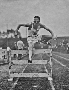A black and white photo captures a male athlete mid-air as he hurdles over an obstacle on a running track, embodying his performance goals. He is wearing a sleeveless top, shorts, and sunglasses. The background shows a blurry crowd and a clear sky.