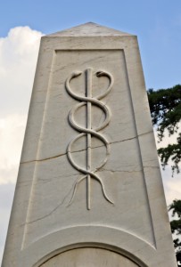 A white stone monument with an engravement of the Rod of Asclepius, symbolizing Health Careers, features a single snake entwined around a staff. The monument is set against a background of a partially cloudy sky and some tree foliage.