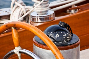 Close-up of a ship's wooden steering wheel beside a compass pointing north, with coiled ropes in the background on a polished wooden deck. The image captures detailed nautical equipment on a sailboat, reminiscent of historic vessels once central to government fleets.