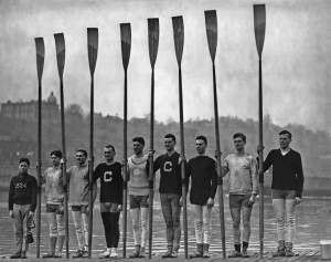 A historic black-and-white photograph shows eight rowers standing side by side on a dock, each holding an oar vertically. Wearing sports uniforms, some display the letter "C" on their chests. A young boy, donning a sweater with "1924" on it, stands at one end—an emblem of teamwork and unity.