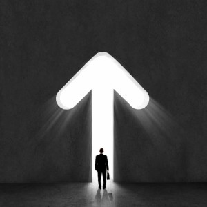 A silhouette of a man holding a briefcase stands before a large illuminated arrow pointing upward on a dark background, suggesting progress, leadership, and direction.