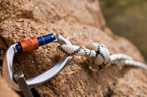 Close-up of a carabiner attached to a rope secured to a rock face, reminiscent of tackling career challenges. The carabiner is metallic with blue and orange accents, and the rope is white with black patterns, tied in a knot. The rock surface appears rugged and textured.