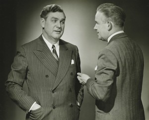 Two men wearing suits engaged in conversation. The man on the left is in a double-breasted suit with a striped pattern and has his hand in his pocket. The man on the right, discussing employee performance, is in a single-breasted suit, facing the other man while gesturing with his hand.