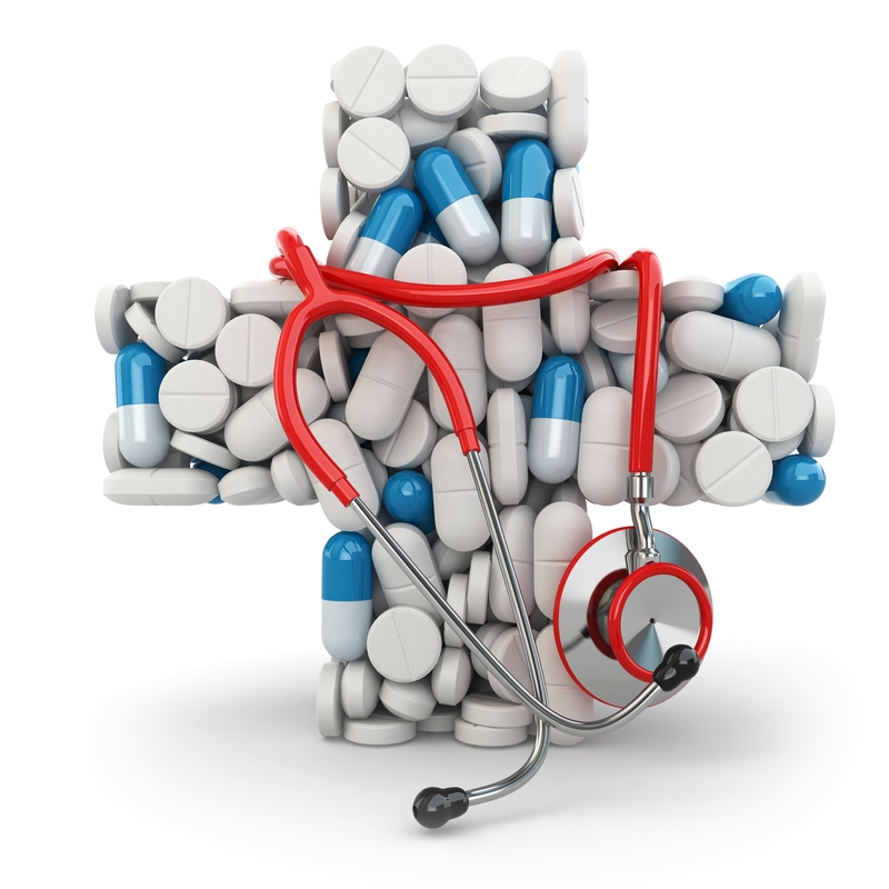 A red stethoscope rests on a cross made of various pills and capsules. The cross, formed by an arrangement of white and blue medication tablets against a white background, symbolizes medical and public health themes.