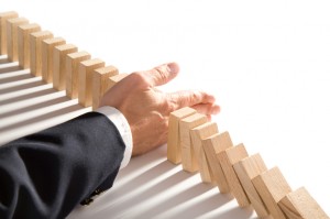 A hand wearing a suit sleeve stops a series of wooden dominoes from toppling in sequence. The dominoes are partially falling, but the hand intervenes to prevent the rest from tumbling over, expertly halting the chain reaction—a perfect metaphor for effective performance management.