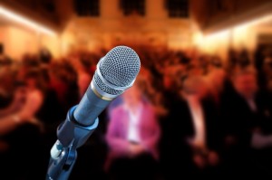 Close-up of a microphone on a stand, centered in the frame. The background shows a blurred public audience in an indoor setting, emphasizing the microphone. The atmosphere suggests a public speaking event, conference, or performance.