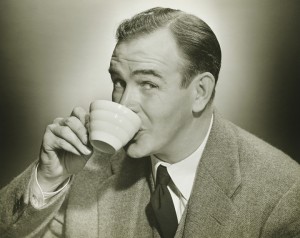 A vintage sepia-toned photograph of a man wearing a suit and tie, taking a break while sipping from a white striped teacup. He is smiling and looking towards the camera. The background is plain and slightly blurred, highlighting the subject.