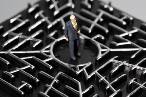 A miniature figurine of a man in a suit stands in the center of a black, metal maze with raised pathways. The man appears to be contemplating his job as he navigates the intricate labyrinth. The image highlights themes of complexity and challenge.
