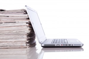 A silver laptop is positioned open beside a large stack of newspapers on a reflective white surface. The laptop screen is blank, and the newspapers are neatly stacked, highlighting the contrast between digital and traditional print media—a scene that could capture any modern employer's workspace.
