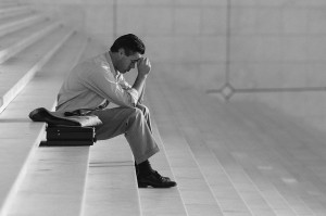 A man in business attire sits on the steps, leaning forward with his elbows resting on his knees and his hands clasped near his face, appearing deep in thought about jobs or career decisions. A briefcase is placed beside him on the step. The background is blurred and minimalistic.