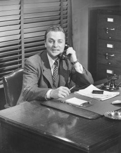 Black and white photo of a man in a suit sitting at a desk with a phone in his hand. He is smiling and looking forward, embodying the optimism of a job seeker. The desk is cluttered with papers, a notepad, and a pen. There are closed blinds behind him and a filing cabinet to his right.