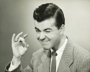 A man with slicked-back hair in a suit and tie is winking and giving an "OK" hand gesture, exuding an aura of trust. The image is in black and white.