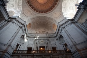 A grand interior view of a neoclassical government building showcases intricate architectural details. The ceiling features ornate carvings and a central dome, while a balcony with decorative railing overlooks a spacious area below. Light streams in through unseen windows above.